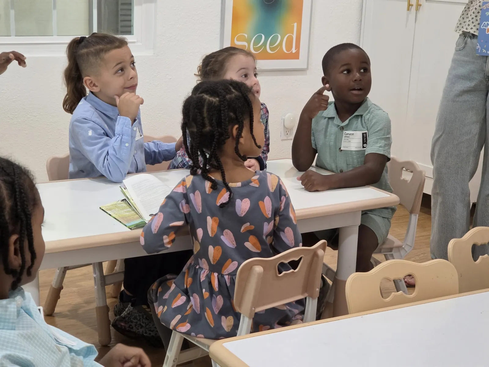 Children seated around a classroom table look toward a teacher during a lesson.