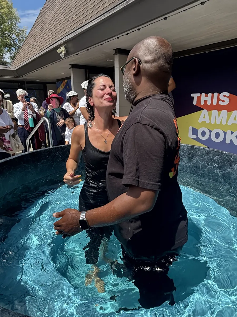 A woman smiles with joy in a baptism pool beside a man, with people watching nearby.
