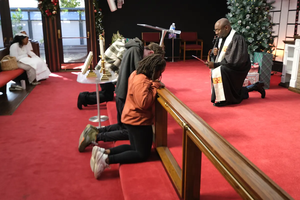 People kneel together at the sanctuary altar rail with others standing nearby.