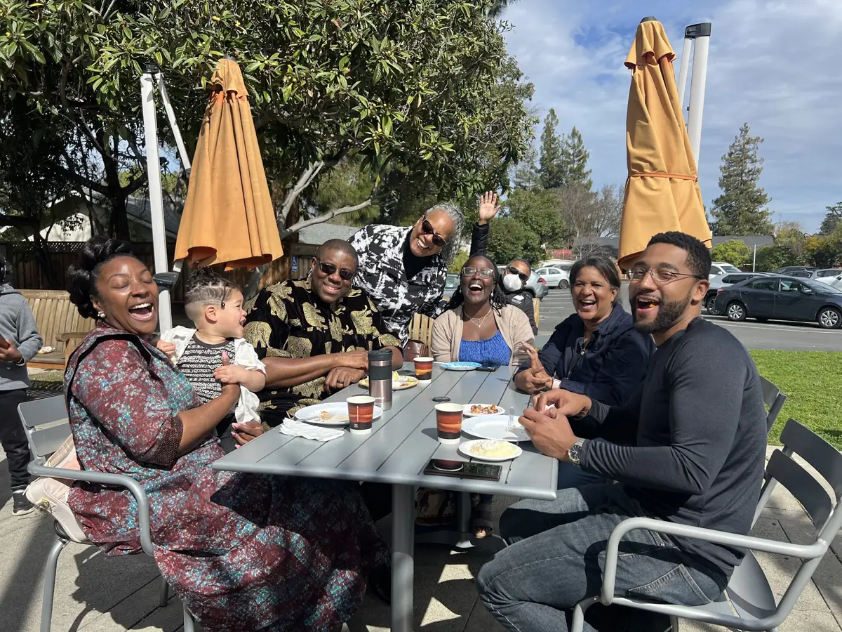 People laugh together around an outdoor table, with an adult holding a child.