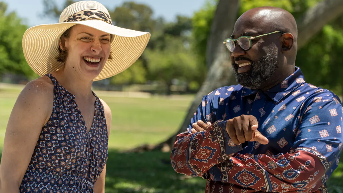 Two people laugh together outdoors beside a picnic table.