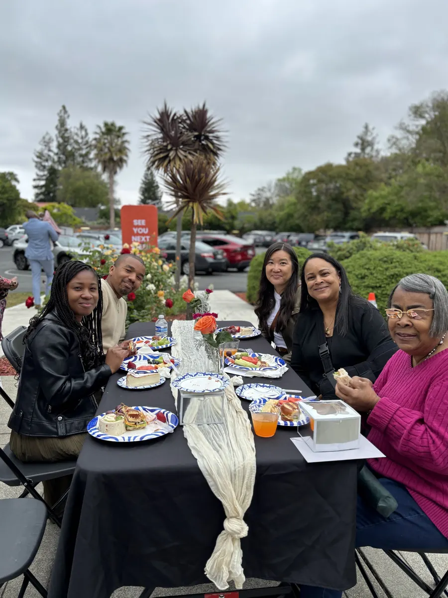 Friends smile together over plates of food at a flower-decorated outdoor table.