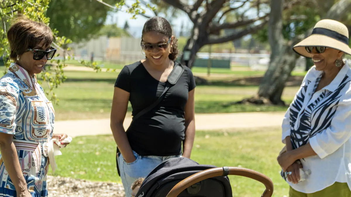 Three people smile and talk together beside a stroller in a leafy park.