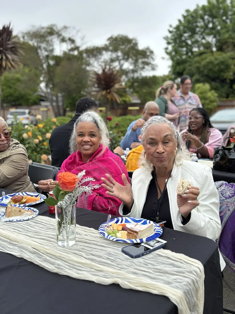 Two people seated at an outdoor lunch table smile with other guests behind them.