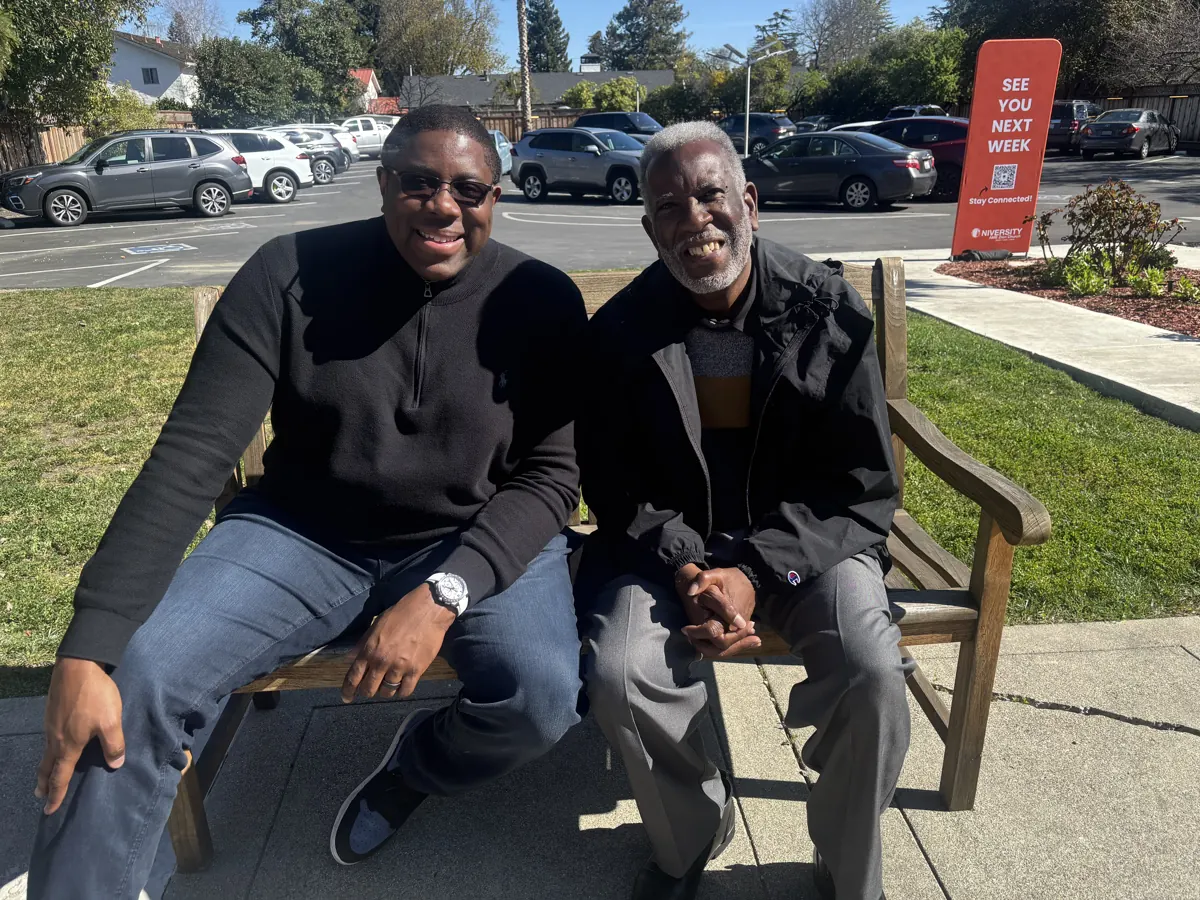 Two men smile together while seated on a bench outside the church.