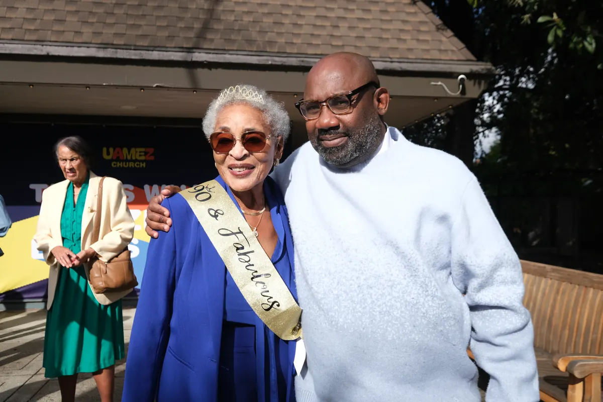 An older woman wearing a sash stands smiling beside a man in the church courtyard.