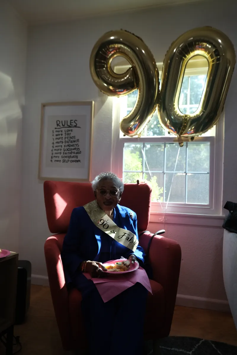 An older woman sits smiling beneath gold number balloons during a birthday celebration.