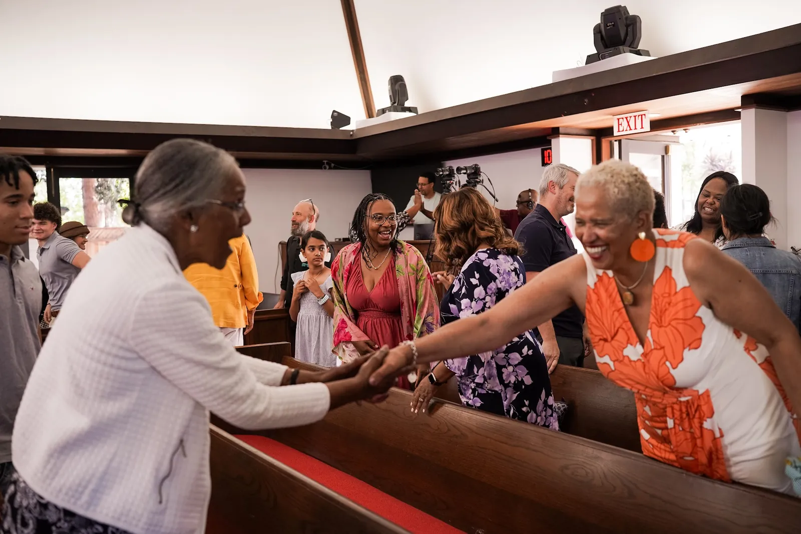 Two people smile and clasp hands across the sanctuary pews while others greet one another.