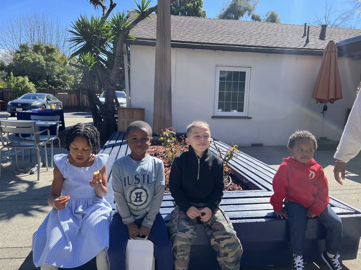 Four children sit together on a bench in the sunny church courtyard.