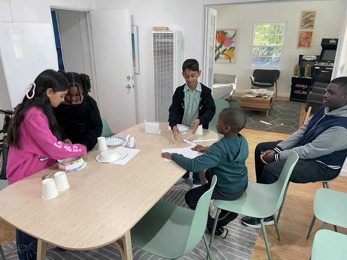 Children gather around a classroom table with craft materials and adults nearby.