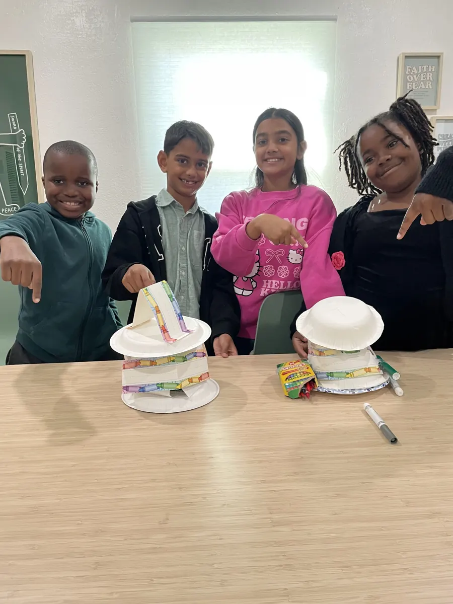 Children and an adult smile behind a classroom table displaying decorated paper hats.