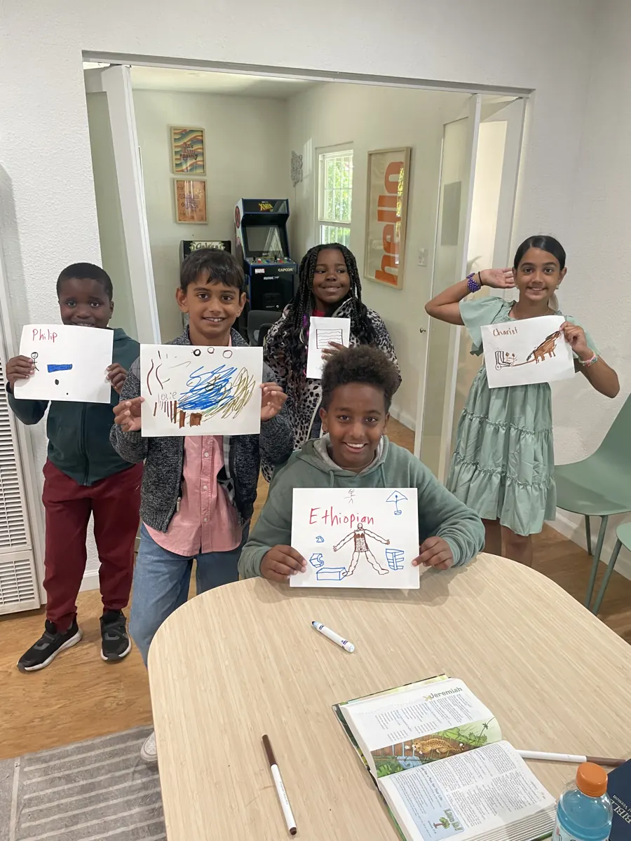 Children stand around a classroom table and proudly hold up their colorful artwork.