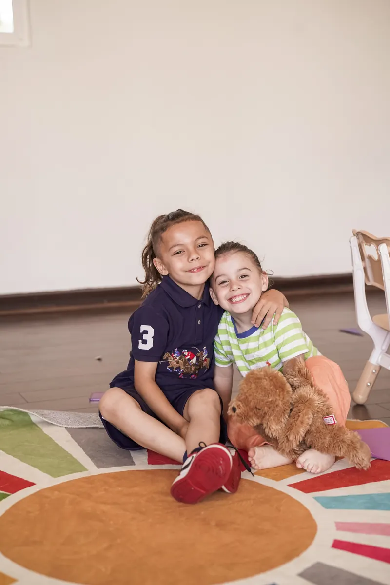 Two children smile together while seated on a colorful classroom rug.
