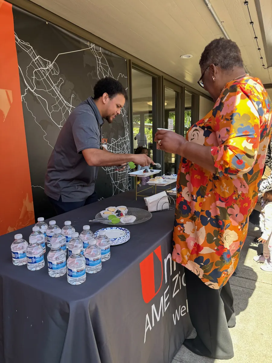 A volunteer greets a guest at an outdoor table with bottled water and printed materials.
