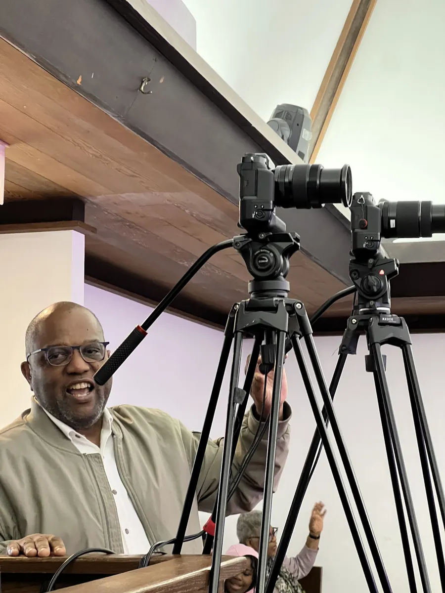 A smiling person sits behind tripod-mounted cameras at the back of the sanctuary.