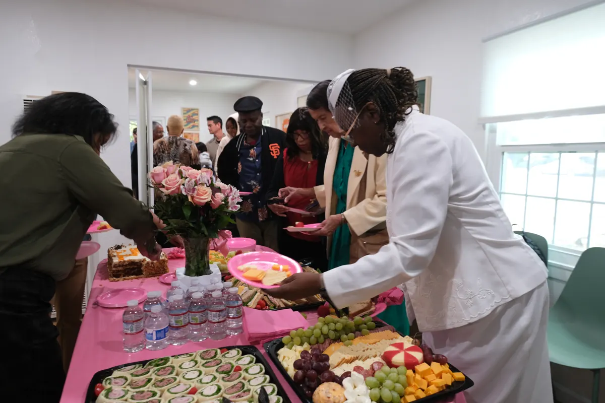 People arrange trays of food and drinks on a table covered with a pink cloth.