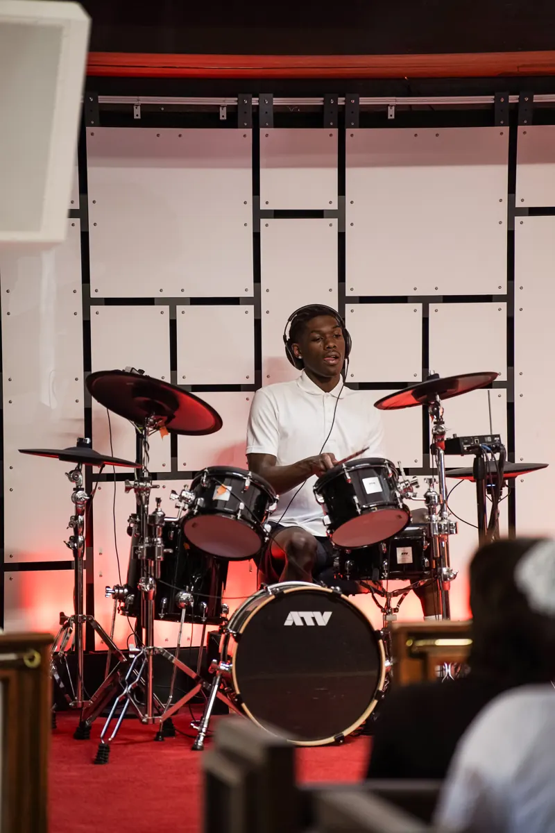 A person in a white shirt plays a drum kit at the front of the sanctuary.