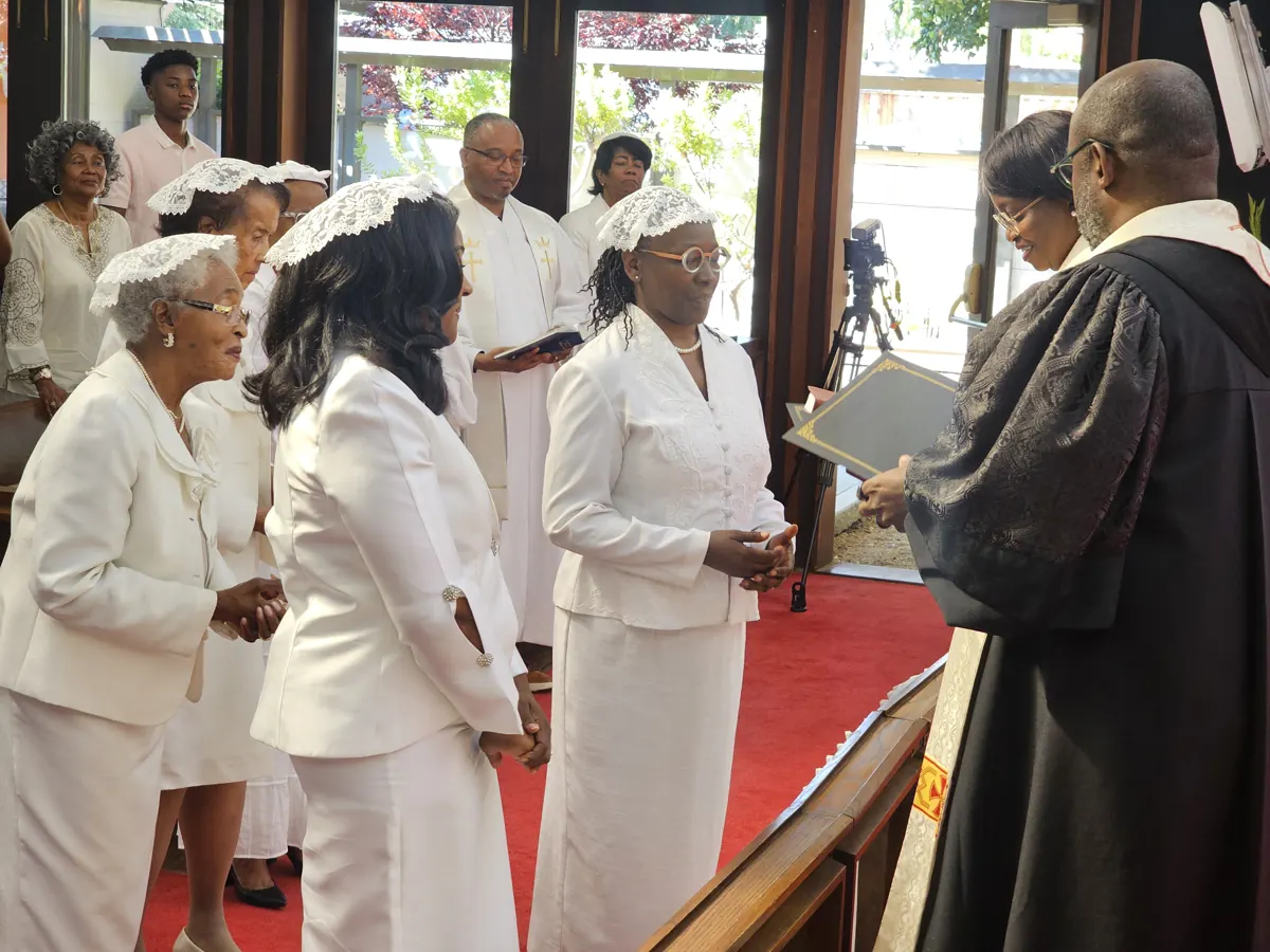 Women dressed in white stand together near the altar rail while a man holds an open book.