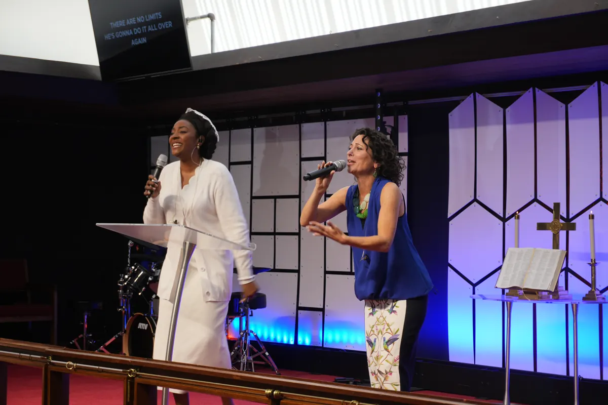 Two women in white and blue sing into microphones on the purple-lit sanctuary stage.