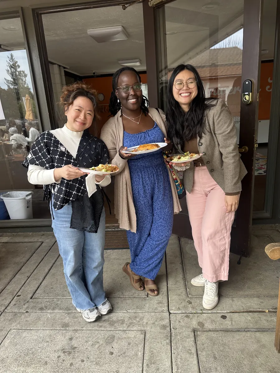 Three friends smile together while holding plates of food outside the church.