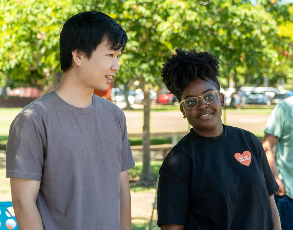 Two young adults smile and talk together beneath the trees at a park.