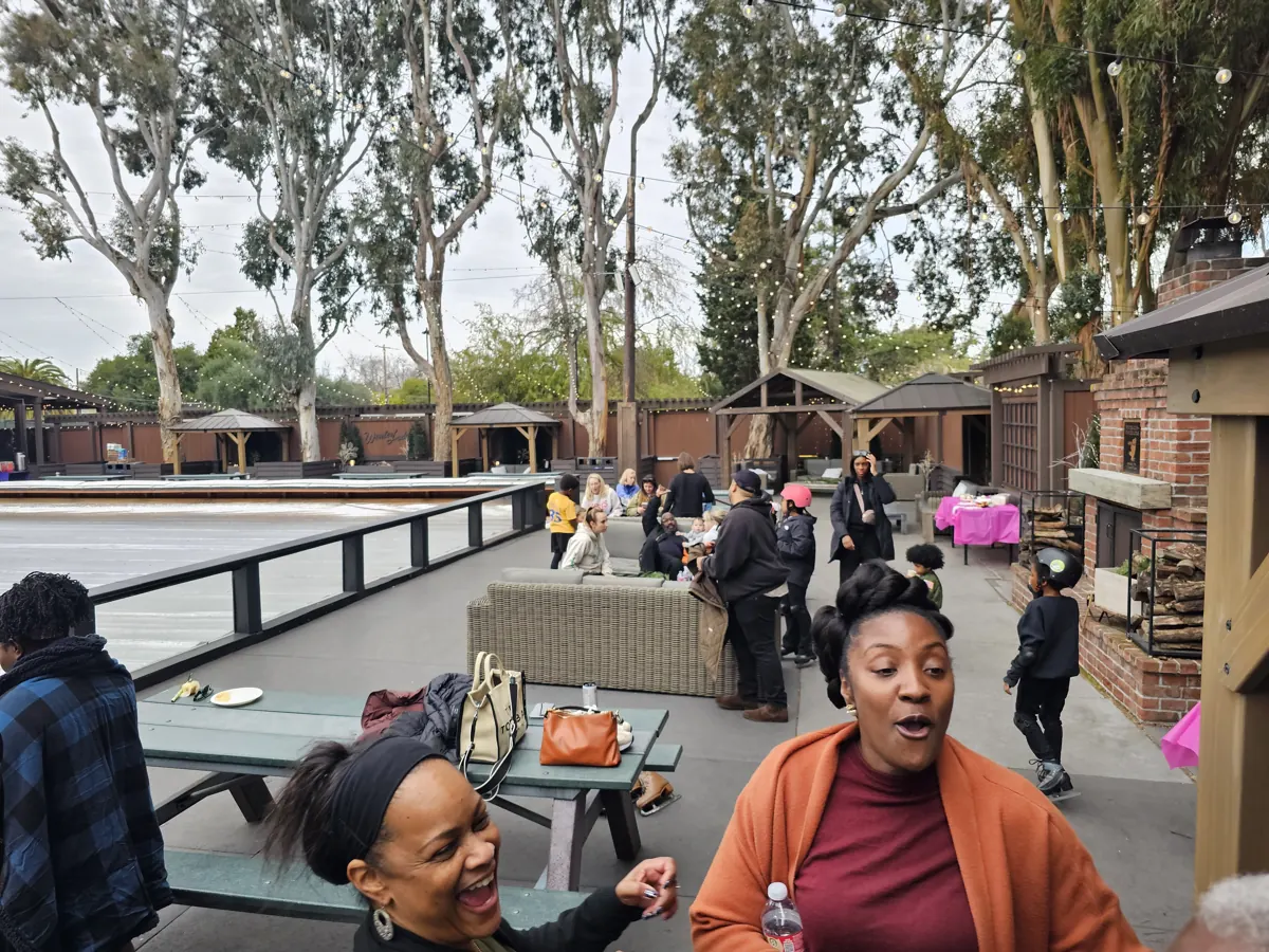 Two friends laugh together beside an outdoor ice rink, with other visitors behind them.